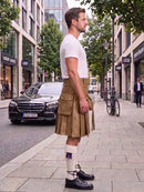 A man standing on a city sidewalk wearing a tan kilt, white t-shirt, white socks, and black shoes, with a street scene and parked cars in the background.