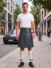 A man stands on a city sidewalk wearing a white crew-neck t-shirt and a dark gray pleated leather kilt, paired with white knee-high socks and black leather shoes. The background shows a street scene with a Mercedes-Benz car, storefronts, and trees.