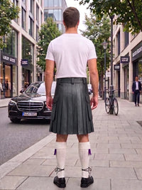 A man viewed from behind wearing a dark gray pleated leather kilt, a white t-shirt, white socks, and black shoes, standing on a city sidewalk.