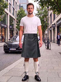 A man stands on a city sidewalk wearing a white short-sleeve t-shirt and a dark gray faux-leather kilt with a matching belt and silver buckle. He is also wearing white knee-high socks and black leather shoes.