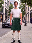 A man stands on a city sidewalk wearing a dark green kilt with silver hardware, a white t-shirt, cream knee-high socks, and black shoes. A black Mercedes-Benz car and urban buildings are visible in the background.
