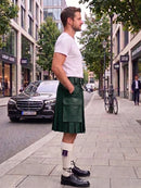 A man standing on a city sidewalk wearing a dark green leather kilt, a white t-shirt, white socks, and black leather shoes. A black Mercedes-Benz car is parked on the street behind him, with trees and storefronts in the background.
