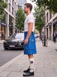 A man standing on a city sidewalk wearing a bright blue leather kilt, a white t-shirt, white socks, and black leather shoes. The background shows a street with a black Mercedes-Benz car, trees, and buildings.