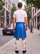 A man standing on a city sidewalk, viewed from behind, wearing a bright blue leather kilt, a white t-shirt, white knee-high socks, and black shoes.