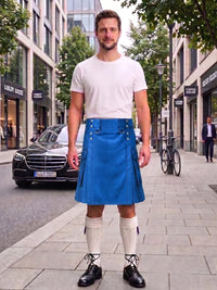 A man stands on a city sidewalk wearing a bright blue kilt with silver metal accents, a white t-shirt, cream knee-high socks, and black leather shoes. A black Mercedes-Benz is parked behind him on the street, with storefronts and trees in the background.