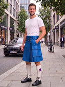 A man standing on a city sidewalk wearing a bright blue leather kilt, a white t-shirt, white knee-high socks, and black leather shoes. The background shows a street with a Mercedes-Benz car, storefronts, and a bicycle.