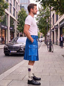 A man standing on a city sidewalk wearing a bright blue pleated kilt, white t-shirt, white knee-high socks, and black leather shoes. The background shows a street with a black Mercedes-Benz car, trees, and storefronts.