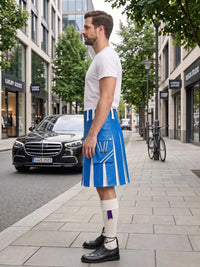 A man standing on a city sidewalk wearing a white t-shirt, a bright blue and white kilt with a front flap pocket, white knee-high socks, and black leather shoes. A black Mercedes-Benz car and a bicycle are parked on the street behind him.
