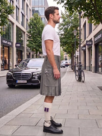 A man standing on a city sidewalk wearing a white t-shirt, a gray pleated kilt with front pockets, white socks with purple stripes, and black leather oxford shoes. A black Mercedes-Benz car and a bicycle are parked on the street behind him.