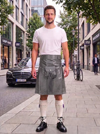 A man stands on a city sidewalk wearing a white short-sleeve t-shirt and a gray pleated leather kilt with silver hardware, paired with white knee-high socks and black leather shoes. Buildings, trees, and a parked black car are visible in the background.