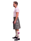 A man shown in profile wearing a gray leather kilt, a light pink t-shirt, white socks with a purple fringed detail, and black leather shoes, set against a plain white background.