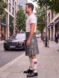 A man standing on a city sidewalk wearing a gray leather kilt, white t-shirt, white socks with purple accents, and black shoes, with a street scene and parked cars in the background.