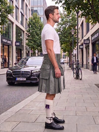 A man standing on a city sidewalk wearing a white short-sleeve t-shirt, a gray pleated kilt with multiple utility pockets, white knee-high socks with a purple accent, and black leather shoes. A black Mercedes-Benz car and a bicycle are visible in the background of the urban street scene.