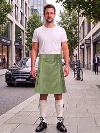 A man stands on a city sidewalk wearing a white crew-neck t-shirt and an olive green pleated kilt with silver button details. He is styled with off-white knee-high socks and black leather shoes, with a street scene featuring a Mercedes-Benz and storefronts in the background.