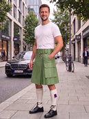 A man wearing a light green kilt with a matching belt, a white t-shirt, white knee-high socks, and black leather shoes, standing on a city sidewalk.