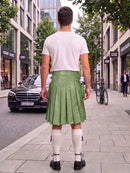 A man standing on a city sidewalk, photographed from behind, wearing a white t-shirt, a green pleated leather kilt, white knee-high socks, and black shoes. A black Mercedes-Benz car is parked on the street to his left, with storefronts and trees in the background.