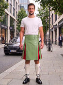 A man stands on a city sidewalk wearing a light green kilt with red side panels, a white t-shirt, white knee-high socks, and black shoes. Buildings, trees, a parked black car, and a bicycle are visible in the background.