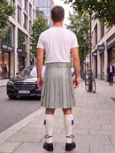 A man viewed from behind wearing a white t-shirt and a pleated light gray kilt, standing on a city sidewalk with a Mercedes-Benz car and storefronts in the background.