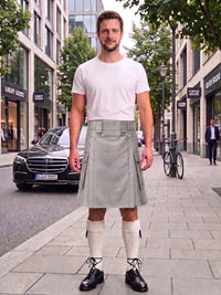 A man stands on a city sidewalk wearing a light gray pleated kilt, a white t-shirt, white knee-high socks, and black leather shoes. The background shows a street with buildings, a parked black Mercedes, and a bicycle.