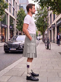 A man standing on a city sidewalk wearing a light gray pleated kilt, white t-shirt, white socks with a purple ribbon, and black leather shoes, with a street scene in the background.
