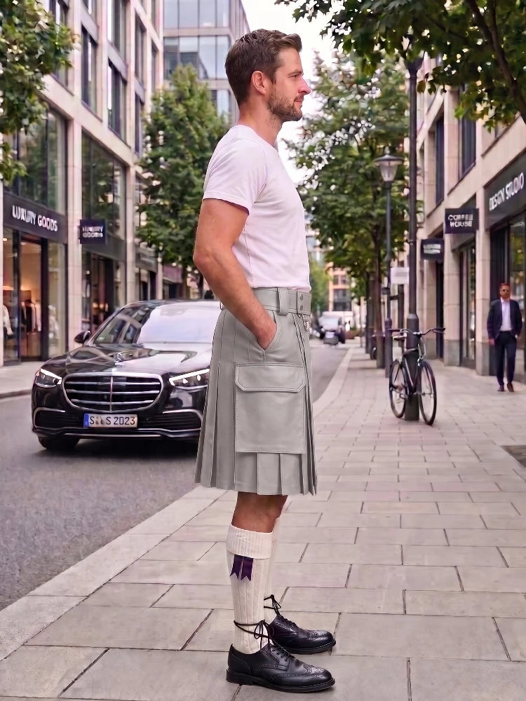 A man standing on a city sidewalk wearing a light gray pleated kilt, white t-shirt, white socks with a purple ribbon, and black leather shoes, with a street scene in the background.