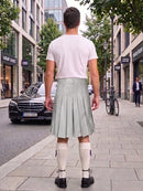 A man standing on a city sidewalk, viewed from behind, wearing a white t-shirt, a pleated light sage green kilt, white knee-high socks, and black shoes. A black Mercedes-Benz car and a bicycle are visible in the street behind him.