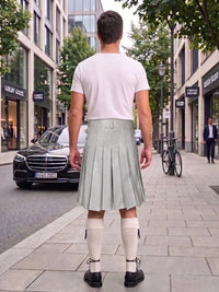 A man standing on a city sidewalk, viewed from behind, wearing a white t-shirt, a pleated light sage green kilt, white knee-high socks, and black shoes. A black Mercedes-Benz car and a bicycle are visible in the street behind him.