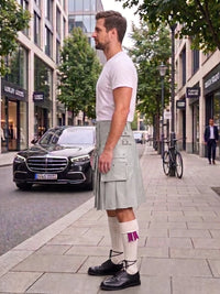 A man standing on a city sidewalk wearing a white t-shirt, a light gray pleated kilt with multiple front pockets, white knee-high socks with purple accents, and black leather shoes. A black Mercedes-Benz car is parked on the street behind him, with trees and storefronts in the background.