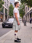 A man standing on a city sidewalk wearing a white short-sleeve t-shirt and a light gray pleated kilt with multiple flap pockets, paired with white knee-high socks and black leather shoes. A black Mercedes-Benz car and a bicycle are visible in the background on a city street.