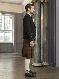 Man wearing a black jacket and plaid kilt standing in an elegant interior setting.