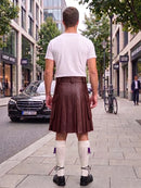 A man standing on a city sidewalk, viewed from behind, wearing a white t-shirt, a dark brown pleated leather kilt, white knee-high socks with purple tassels, and black shoes.