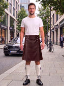 A man stands on a city sidewalk wearing a dark green kilt with silver hardware, a white t-shirt, cream knee-high socks, and black shoes. A black Mercedes-Benz car and urban buildings are visible in the background.