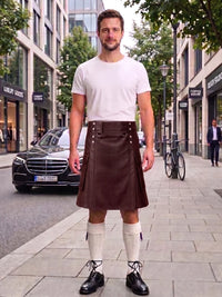 A man stands on a city sidewalk wearing a dark green kilt with silver hardware, a white t-shirt, cream knee-high socks, and black shoes. A black Mercedes-Benz car and urban buildings are visible in the background.