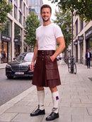 A man stands on a city sidewalk wearing a dark brown leather kilt, a white t-shirt, white knee-high socks, and black leather shoes. The background shows a street with buildings, a parked black Mercedes, and a bicycle.