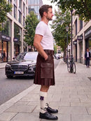 A man standing on a city sidewalk wearing a dark green pleated kilt, a white t-shirt, white socks, and black leather shoes. A black Mercedes-Benz car is parked on the street behind him, with trees and storefronts in the background.