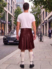 A man standing on a city sidewalk, viewed from behind, wearing a white t-shirt and a dark brown pleated faux-leather kilt with white knee-high socks and black shoes.