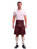 A man modeling a black faux-leather kilt with a matching black waist belt and pouch, paired with a white t-shirt, cream knee-high socks, and black lace-up shoes, against a plain white background.