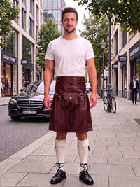 A man stands on a city sidewalk wearing a white crew-neck t-shirt and a dark brown leather kilt with a front pouch and strap details, paired with white knee-high socks and black leather shoes.