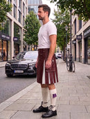 A man stands on a city sidewalk wearing a white t-shirt, a dark brown leather kilt with white pleated inserts, white knee-high socks with a purple bow, and black leather shoes. A black Mercedes-Benz car and storefronts are visible in the background.