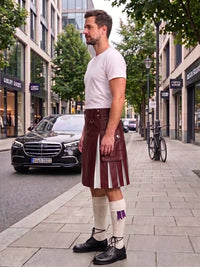 A man stands on a city sidewalk wearing a white t-shirt, a dark brown leather kilt with white pleated inserts, white knee-high socks with a purple bow, and black leather shoes. A black Mercedes-Benz car and storefronts are visible in the background.