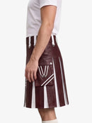 A person wearing a dark burgundy faux-leather kilt with bold white vertical stripes and silver-tone hardware, paired with a white t-shirt against a light gray background.