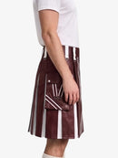 A side-profile view of a person wearing a pleated kilt in a deep burgundy faux leather with bold white vertical stripes and white drawstrings, paired with a white t-shirt against a plain white background.