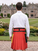 A man viewed from behind wearing a white dress shirt and a bright red pleated kilt with black waist belt and black side pockets, standing in a garden with a large stone mansion in the background.
