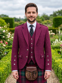A man wearing a traditional Scottish formal outfit with a burgundy double-breasted jacket, matching vest, tartan kilt, and a brown sporran, standing in a garden setting.