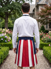 A man viewed from behind wearing a white long-sleeve dress shirt and a pleated kilt with vertical red, white, and navy blue stripes, standing in a garden path.