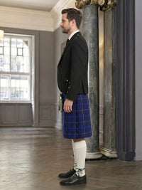 Man wearing a dark suit and blue plaid kilt in an elegant interior setting.