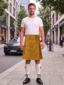 A man stands on a city sidewalk wearing a white crew-neck t-shirt and a mustard yellow corduroy kilt, paired with white knee-high socks and black leather shoes. The background shows a street scene with a black Mercedes-Benz, trees, and storefronts.