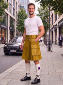 A man stands on a city sidewalk wearing a mustard yellow kilt with a matching belt and buckle, a white t-shirt, white knee-high socks, and black shoes. A black Mercedes-Benz car is parked on the street behind him, with trees and storefronts in the background.