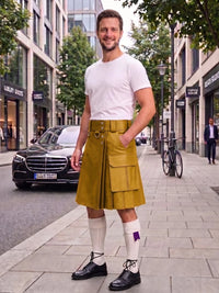 A man stands on a city sidewalk wearing a mustard yellow kilt with a matching belt and buckle, a white t-shirt, white knee-high socks, and black shoes. A black Mercedes-Benz car is parked on the street behind him, with trees and storefronts in the background.
