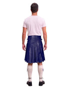 A man shown from the back wearing a dark blue pleated kilt, a light pink t-shirt, cream-colored knee-high socks, and black shoes, against a solid white background.